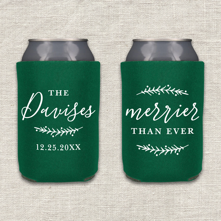 Two collapsible foam can coolers are shown on a textured beige background. The can coolers are forest green, and the writing on the can coolers is white. The can cooler on the left says the couple's last name and wedding date, along with a pine branch. The can cooler on the right says "Merrier Than Ever," along with two pine branches.
