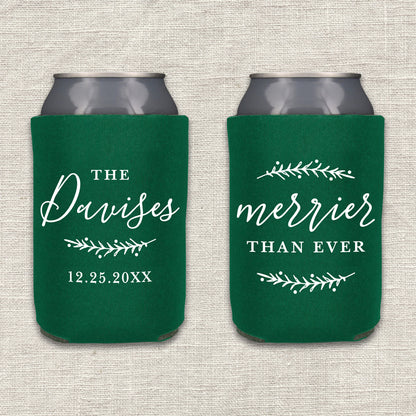 Two collapsible foam can coolers are shown on a textured beige background. The can coolers are forest green, and the writing on the can coolers is white. The can cooler on the left says the couple's last name and wedding date, along with a pine branch. The can cooler on the right says "Merrier Than Ever," along with two pine branches.