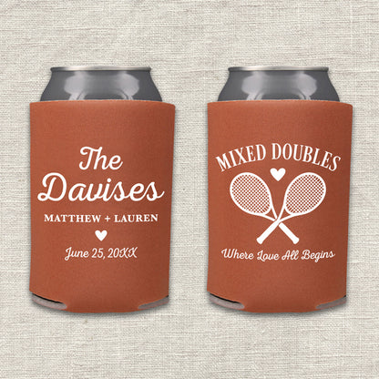 Two rust-colored can coolers displayed side by side on a light beige textured fabric background. The left can cooler is personalized with 'The Davises', the names 'Matthew + Lauren', and a wedding date. The right can cooler features the text 'Mixed Doubles - Where Love All Begins.'