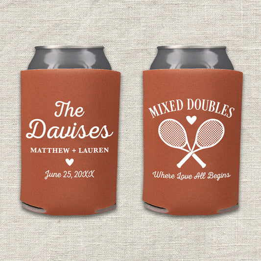 Two rust-colored can coolers displayed side by side on a light beige textured fabric background. The left can cooler is personalized with 'The Davises', the names 'Matthew + Lauren', and a wedding date. The right can cooler features the text 'Mixed Doubles - Where Love All Begins.'