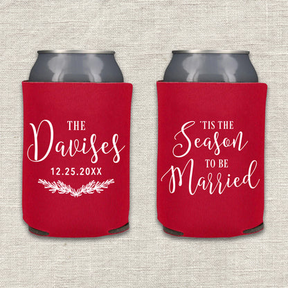 Two collapsible foam can coolers are shown on a textured beige background. The can coolers are red, and the writing on the can coolers is white. The can cooler on the left says the couple's last name and wedding date, along with a pine branch laurel. The can cooler on the right says "'Tis the Season to be Married."